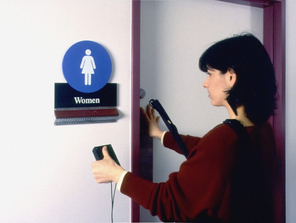 Frau vor Tür mit Aufschrift "Women" und Braille-Reader sowie Langstock in der Hand